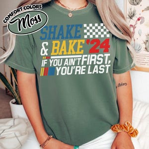 May include: A moss green t-shirt with the words "SHAKE & BAKE '24" in blue, yellow, and red. The shirt also features the phrase "IF YOU AIN'T FIRST, YOU'RE LAST" in white. A checkered flag design is above the text.