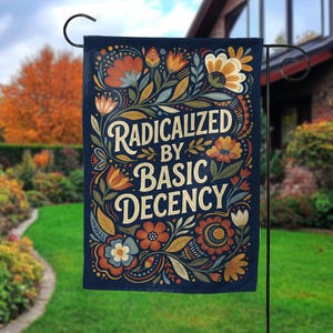 May include: A navy blue garden flag featuring the text "Radicalized by Basic Decency" in a decorative font, framed by orange, yellow, and blue floral designs. The flag is displayed in a garden setting.