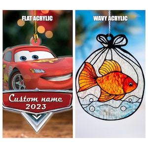 K&ouml;nnte beinhalten: Zwei Weihnachtsornamente aus Acryl. Eines zeigt einen roten Cartoon-Rennwagen mit dem Text "Custom name 2023". Das andere ist ein wellenf&ouml;rmiges Acrylornament mit einem orangefarbenen Goldfisch-Design.