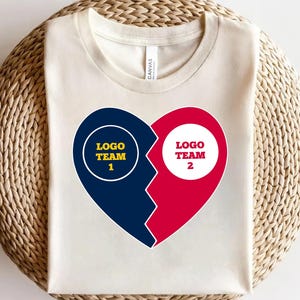 May include: Cream-colored t-shirt featuring a split heart design. One half is navy blue with "LOGO TEAM 1" in a yellow circle, the other half is red with "LOGO TEAM 2" in a white circle. The shirt is made of a soft material.