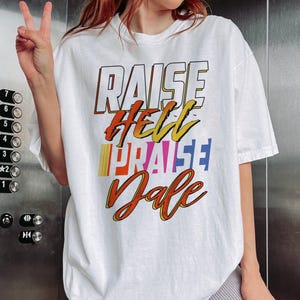 May include: White t-shirt with the text "RAISE HELL PRAISE Dale" in a colorful, retro font. The text is layered with orange, yellow, blue, and red accents. The shirt has short sleeves and a relaxed fit.