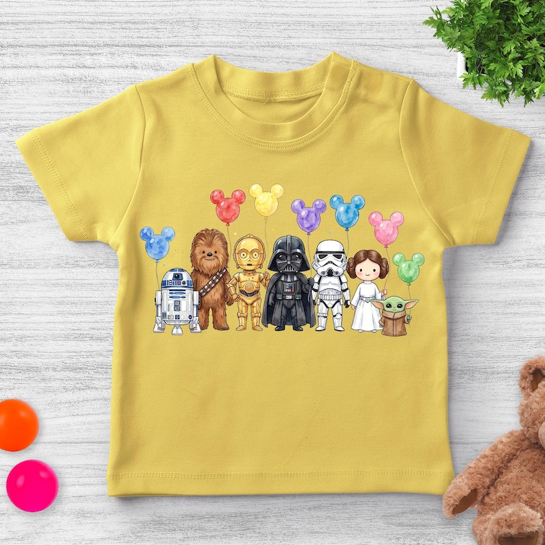 Puede incluir: Camiseta amarilla de manga corta con una colorida ilustraci&oacute;n de personajes de Star Wars sosteniendo globos con forma de Mickey Mouse. Los personajes incluyen a R2-D2, Chewbacca, C-3PO, Darth Vader, un Stormtrooper, la Princesa Leia y Grogu.