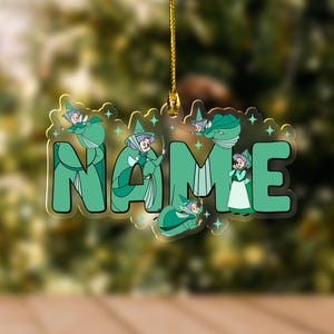 May include: A clear acrylic ornament with the word "NAME" in green, featuring cartoon fairy illustrations. The ornament is suspended by a gold string, with a blurred green and brown background.