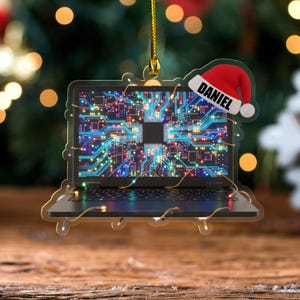 K&ouml;nnte beinhalten: Ein weihnachtlicher Laptop-Ornament mit einem Leiterplatten-Design auf dem Bildschirm. Das Ornament hat einen Weihnachtsmannhut mit dem Namen "DANIEL". Es ist an einer goldenen Schnur aufgeh&auml;ngt und wirkt festlich.