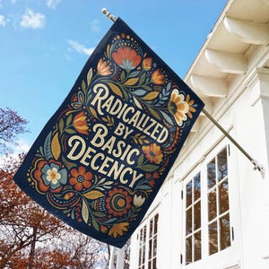 May include: A navy blue flag displaying the words "Radicalized by Basic Decency" in a decorative font, surrounded by orange, yellow, and blue floral and leaf patterns. The flag is attached to a white building, set against a blue sky.