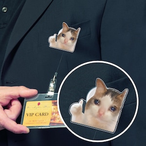 May include: A badge featuring a meme of a cat with a sad expression and a thumbs-up gesture. The badge is attached to a retractable lanyard and is clipped to a dark blue blazer. A VIP card is also visible.