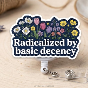 May include: A white and navy blue badge reel with the text "Radicalized by basic decency" and a floral design. The flowers include daisies, tulips, and other colorful blooms. The reel is on a wooden surface.