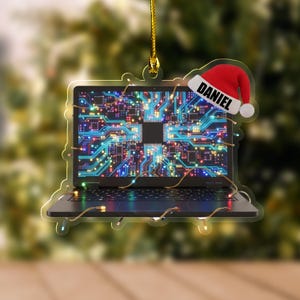 K&ouml;nnte beinhalten: Ein weihnachtlicher Laptop-Ornament mit einem Leiterplattendesign auf dem Bildschirm. Der Schmuck hat einen Weihnachtsmannhut mit dem Namen "DANIEL". Das Ornament ist mit kleinen Lichtern und einem goldenen Faden zum Aufh&auml;ngen verziert.