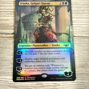 May include: A holographic Magic: The Gathering card featuring Vraska, Golgari Queen. The card has a green, blue, and purple color scheme with a detailed illustration of a woman with snakes for hair. The card text describes abilities and game mechanics.