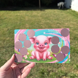 May include: A colorful scratch-off card featuring a cartoon pig with a cheerful expression, set against a backdrop of pastel colors and butterflies. The card has ten scratch-off circles and the text "$50 1-10" and "Brits Budgets Life With Me".
