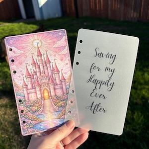 May include: A pink and purple planner insert with a castle illustration and the text "Saving for my Happily Ever After". The insert is held in front of a green background.