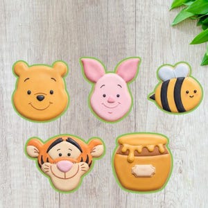 May include: A collection of five character-shaped cookies. The cookies include Winnie the Pooh, Piglet, a bee, Tigger, and a honey pot. Each cookie has a green border and is set against a light wood-grain background.