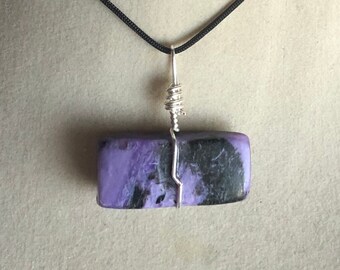 May include: A purple and black stone pendant with silver wire wrapping. The pendant is hanging from a black cord.