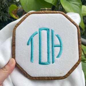 K&ouml;nnte beinhalten: Gesticktes Monogramm in t&uuml;rkisfarbenem Garn auf wei&szlig;em Stoff, eingefasst in einen achteckigen Holz-Stickrahmen. Der Holzrahmen hat ein dekoratives Holzmaserungsmuster. Das Monogramm scheint die Buchstaben TDA zu sein.