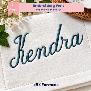 May include: A white fabric square featuring the dark teal embroidered name Kendra in a cursive script. A pink banner at the top displays the text Embroidery Font 6 Sizes 2.54cm 3.81cm 5.08cm 6.35cm 7.62cm 8.89cm and the bottom text reads +BX Formats. The design is accented by white flowers.