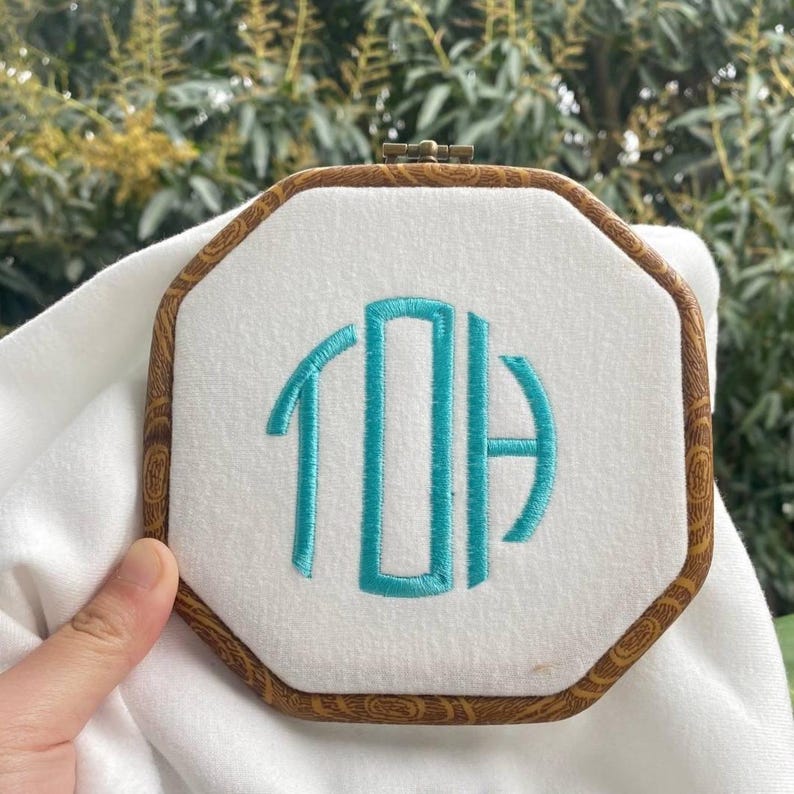 K&ouml;nnte beinhalten: Ein achteckiger Stickrahmen mit einem wei&szlig;en Stoffmittelteil mit t&uuml;rkisfarbenen Monogrammbuchstaben. Der Holzrahmen hat ein dekoratives Holzmaserungsmuster und einen Metallverschluss. Der Hintergrund ist unscharf.