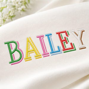 May include: A white fabric garment featuring the embroidered name BAILEY in a multi-colored block font. Each letter is stitched in a different color, including green, yellow, pink, blue, red, and brown, with a layered outline effect.