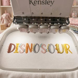 May include: A Kensley embroidery machine with a white fabric hoop featuring the embroidered text DISNOSOUR in multi-colored, dinosaur-themed letters. The machine has numbered needle positions from 2 to 6 visible above the white fabric surface.