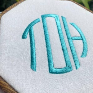 K&ouml;nnte beinhalten: Nahaufnahme eines wei&szlig;en Stoffes mit einer t&uuml;rkisfarbenen Monogramm-Stickerei in einem h&ouml;lzernen Stickrahmen. Das Monogramm zeigt die Buchstaben TDA in einem stilisierten, verbundenen Design. Das Stickgarn hat eine leuchtende t&uuml;rkise Farbe.