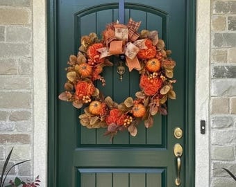 May include: A fall-themed wreath with orange pumpkins, flowers, and leaves. The wreath is adorned with a large bow and hangs on a dark green door. The door has gold hardware and is set in a stone facade.