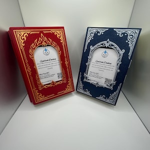 May include: Two rectangular deck boxes shaped like books, one red with gold filigree and one blue with silver filigree. Each features a central arched window displaying a white card with the text Spellbook Deckbox, a logo, and a QR code.