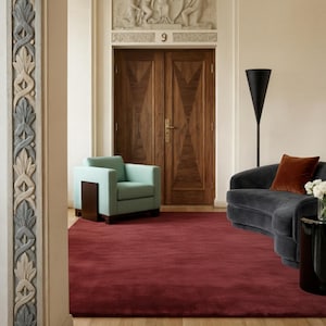 Peut inclure: Intérieur d'un salon avec un tapis bordeaux, un fauteuil turquoise et un canapé incurvé gris foncé avec un coussin rouille. La pièce comprend des portes en bois et un lampadaire noir, créant une atmosphère moderne et élégante.