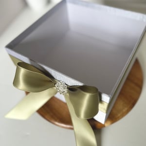 May include: A white gift box with a satin ribbon tied in a bow with a rhinestone embellishment. The box is sitting on a wooden surface.
