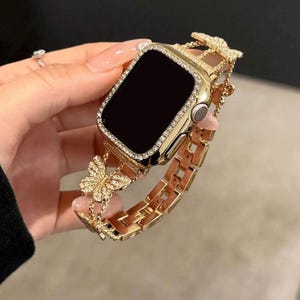 May include: Gold-toned Apple Watch with a decorative band featuring butterfly accents. The watch case is encrusted with small, clear crystals. The band is made of gold-colored metal links and butterfly charms.