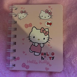 May include: A pink Hello Kitty notebook with a spiral binding. The cover features Hello Kitty in different poses with pink bows and hearts.