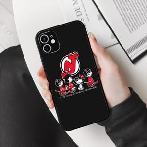 May include: Black phone case with a white outline of the New Jersey Devils logo and five Peanuts characters wearing hockey jerseys. The characters are holding hockey sticks and a puck.