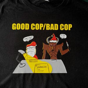 Puede incluir: Camiseta negra con un gráfico humorístico. El texto "GOOD COP/BAD COP" es amarillo. El diseño presenta a Papá Noel y Krampus, con bocadillos de diálogo y una lista de traviesos. La lista incluye "You" y "Your Mom".