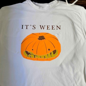 Puede incluir: Camiseta blanca con el texto "IT'S WEEN" y un gran diseño de calabaza naranja. La calabaza tiene un tallo marrón y una boca verde y marrón. La camiseta está sobre una superficie oscura.