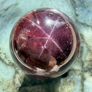 Puede incluir: Una gema esférica transparente con un tono burdeos intenso. La superficie pulida refleja la luz, revelando un patrón en forma de estrella. La gema se encuentra sobre un fondo moteado de color azul verdoso claro.