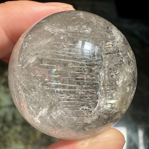 Peut inclure: Une sphère de cristal de quartz clair, présentant des fractures et inclusions internes. La surface polie reflète la lumière, créant un effet chatoyant. La sphère est tenue dans une main, soulignant sa taille et sa clarté. Un objet décoratif.