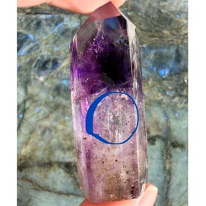 Puede incluir: Una punta de cristal de amatista pulida, que muestra un degradado de colores desde el púrpura intenso hasta el lavanda translúcido. El cristal es sostenido por una mano, revelando su belleza natural. Las facetas de la piedra capturan la luz, resaltando sus patrones internos.
