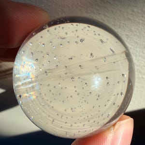 May include: A transparent glass sphere, possibly a paperweight, filled with small air bubbles. The orb is held by fingers, and light passing through creates a rainbow effect. The design is simple and elegant, with a focus on clarity and light refraction.