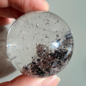 May include: A clear quartz crystal sphere with dark inclusions. The sphere is held in a hand and appears polished. The inclusions are a mix of black and brown, creating a unique pattern within the crystal.