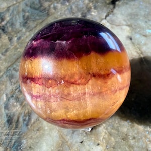 Puede incluir: Una gema esférica pulida con bandas de color. La parte superior es de color púrpura oscuro, que pasa a naranja y luego a un amarillo anaranjado más claro en la base. Venas rojas recorren las capas naranjas. La esfera es lisa y reflectante.