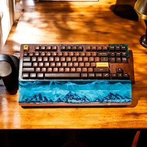 Puede incluir: Un teclado marrón y negro con un reposamuñecas de resina azul y blanco. El teclado tiene una variedad de teclas, incluidas teclas de función, teclas de flecha y teclas numéricas. El reposamuñecas tiene un diseño de cordillera.