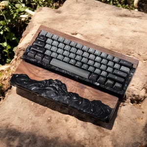 May include: A custom mechanical keyboard with a wooden frame and dark grey keycaps. The wrist rest has a black resin design. The keyboard has a standard QWERTY layout. The keyboard is resting on a stone surface.