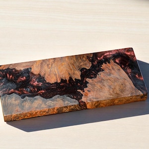 May include: A rectangular wooden serving board with a unique design. The board features a combination of natural wood tones and a dark red, almost black, resin. The wood grain is visible, adding to the rustic charm.
