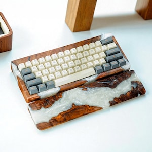 Pode incluir: Um teclado mecânico personalizado com estrutura de madeira e um conjunto de teclas brancas e cinzentas. A estrutura é incrustada com um design de resina branca e cinzenta. Uma almofada de pulso a condizer também está incluída.