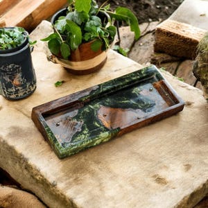 May include: A rectangular wooden tray with a dark green and brown marbled resin inlay. The tray has a raised edge and small metal accents. The tray is on a stone surface, with potted plants in the background.