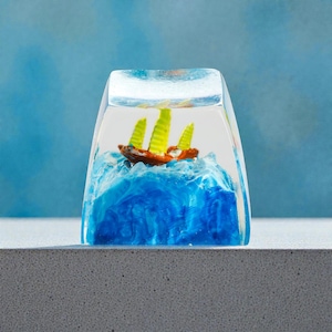 May include: A clear resin block encases a miniature sailboat on a blue ocean. The boat is brown with yellow sails, set against a backdrop of swirling blue resin. The base is a light gray surface.