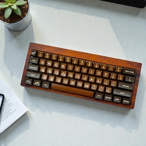 May include: A wooden keyboard with brown keycaps. The keyboard has a compact layout with various function keys and alphanumeric keys. The keys are labeled with white lettering. The keyboard is on a light-coloured surface, with a small plant in a pot visible.