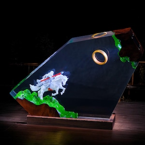 Può includere: Scultura decorativa in resina raffigurante una figura bianca a cavallo con una lancia rossa, su sfondo scuro. La scultura presenta un anello dorato e dettagli verdi, montata su una base di legno.