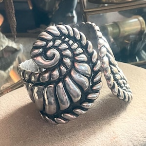 May include: A silver-toned ring with a spiral design. The ring features a textured, braided band that wraps around the finger, with a detailed, shell-like spiral at the center. The ring is made of a shiny, metallic material.