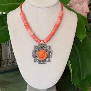May include: A coral-colored beaded necklace with a silver pendant. The pendant features an orange rose in the center, surrounded by an ornate silver design. The necklace is displayed on a mannequin.