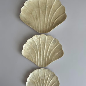 May include: A set of three cream-colored, shell-shaped dishes arranged vertically. Each dish features a scalloped edge and a textured, ribbed surface that mimics the natural appearance of a seashell, with a glossy finish reflecting light.