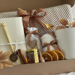 May include: A gift box filled with a white mug, a notebook, a pen, a blanket, a jar of honey, a honey dipper, dried orange slices, and a small bag tied with a ribbon. The box is brown and the items are arranged inside.